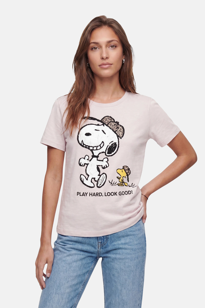Eine Frau mit langen braunen Haaren trägt ein hellrosa „Frogbox by Princess Limited Edition Snoopy“-T-Shirt aus weicher Baumwolle, auf dem Snoopy und Woodstock mit Hüten und dem Schriftzug „PLAY HARD, LOOK GOOD!“ abgebildet sind. Vor einem einfarbigen Hintergrund posiert sie mit einer Hand in der Hüfte.