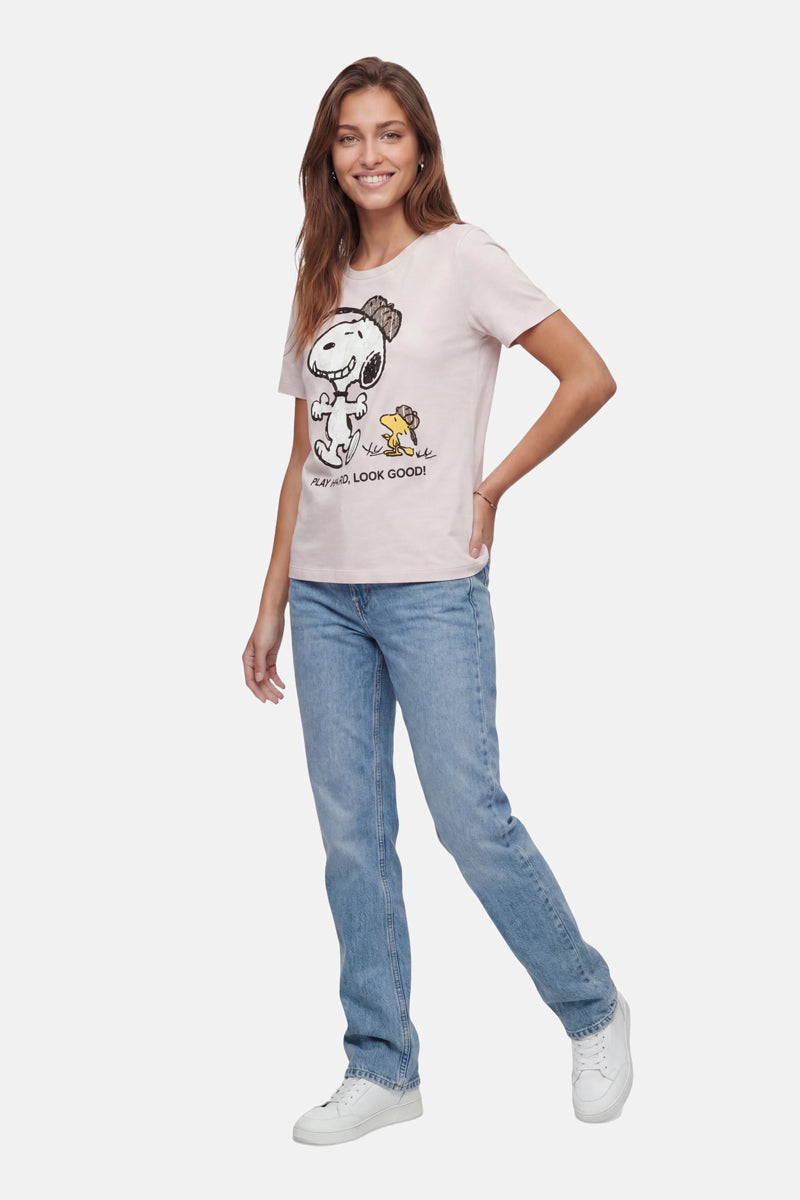 Eine Frau mit langen braunen Haaren trägt ein „Frogbox by Princess Limited Edition Snoopy“-T-Shirt aus Baumwolle, hellblaue Jeans und weiße Sneakers; sie lächelt mit einer Hand in die Hüfte gestützt vor einem schlichten weißen Hintergrund.