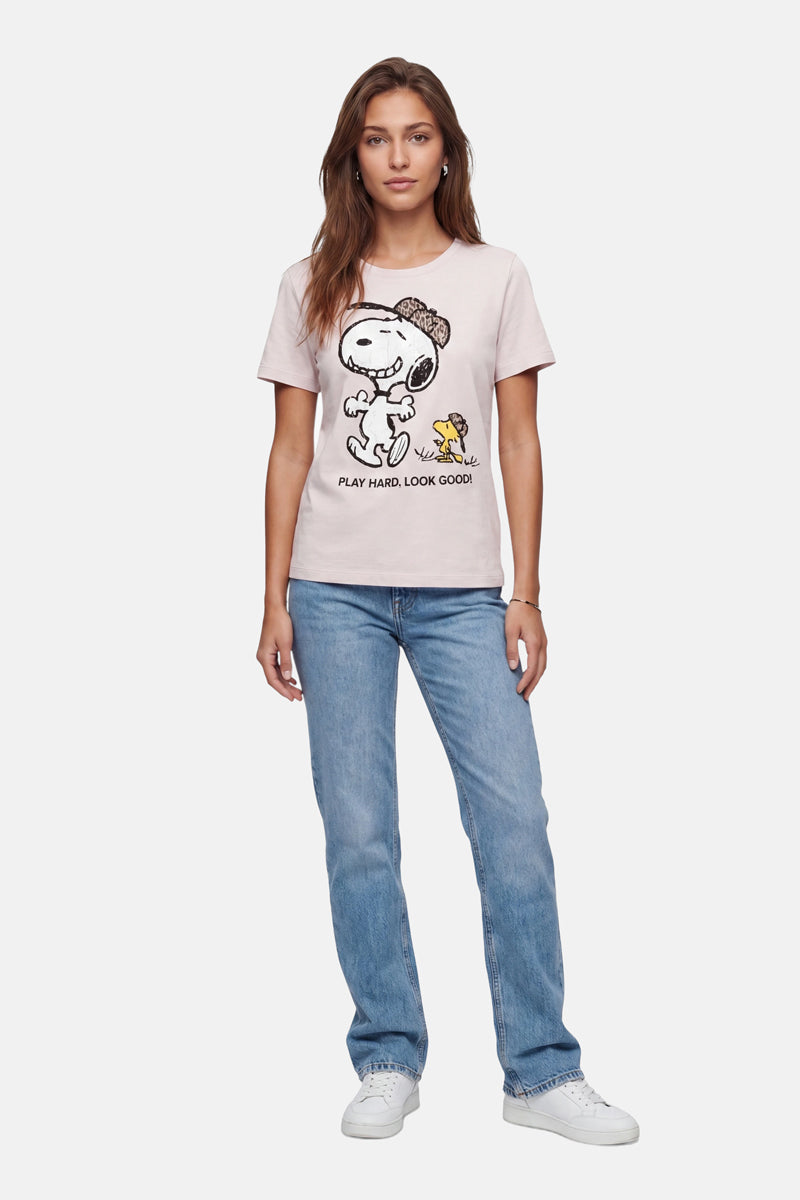 Eine Frau mit langen braunen Haaren trägt ein hellrosa Snoopy-T-Shirt aus Baumwolle der „Frogbox by Princess Limited Edition“ mit der Aufschrift „Play Hard Look Good!“, hellblaue Jeans und weiße Sneakers und steht entspannt vor einem schlichten weißen Hintergrund.