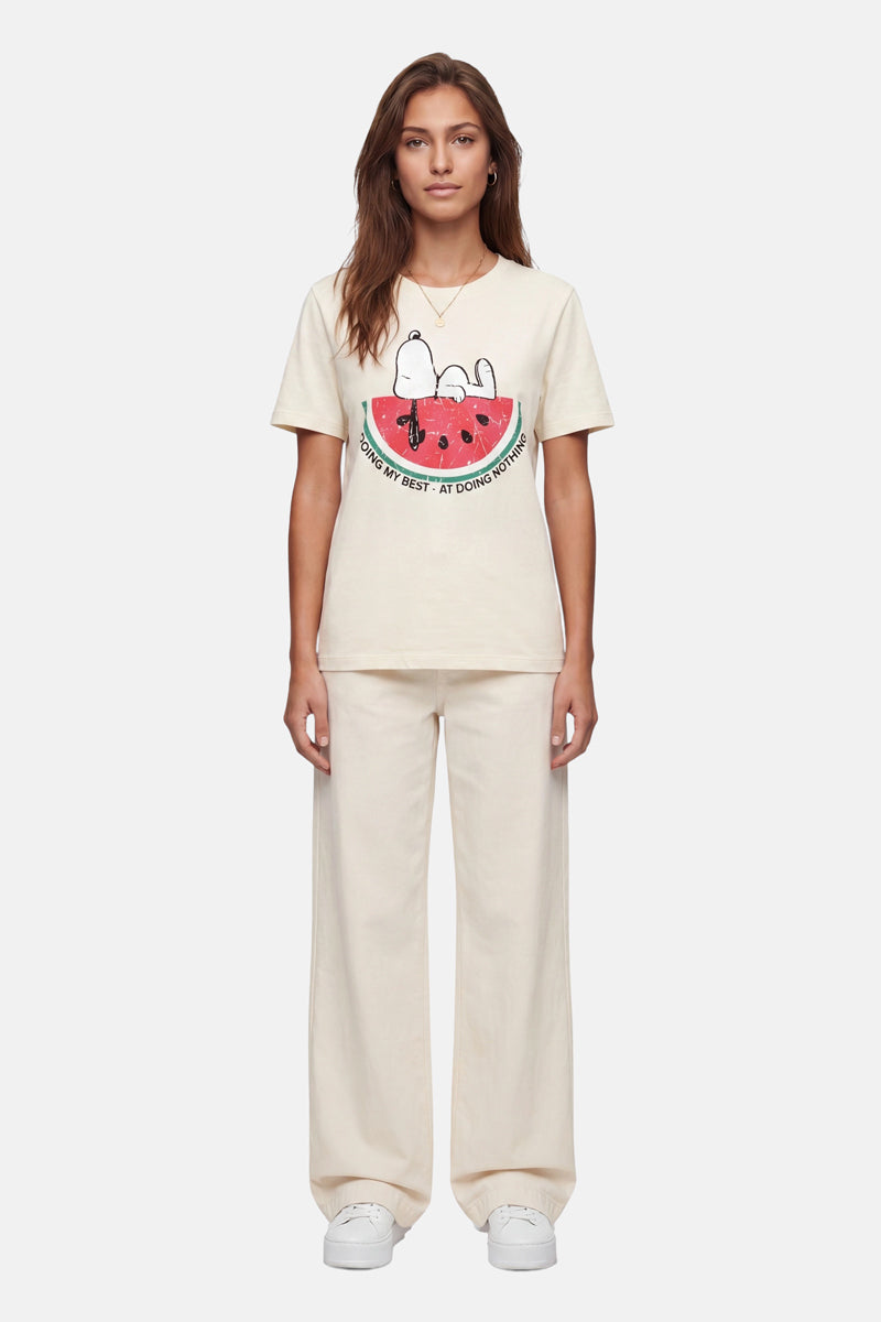 Eine Frau blickt nach vorne in einem cremefarbenen „Frogbox by Princess Snoopy“-T-Shirt aus Baumwolle, auf dem Snoopy und Woodstock auf einer Wassermelone zu sehen sind. Dazu trägt sie eine passende Hose mit weitem Bein und weiße Plateau-Sneaker vor einem schlichten weißen Hintergrund.