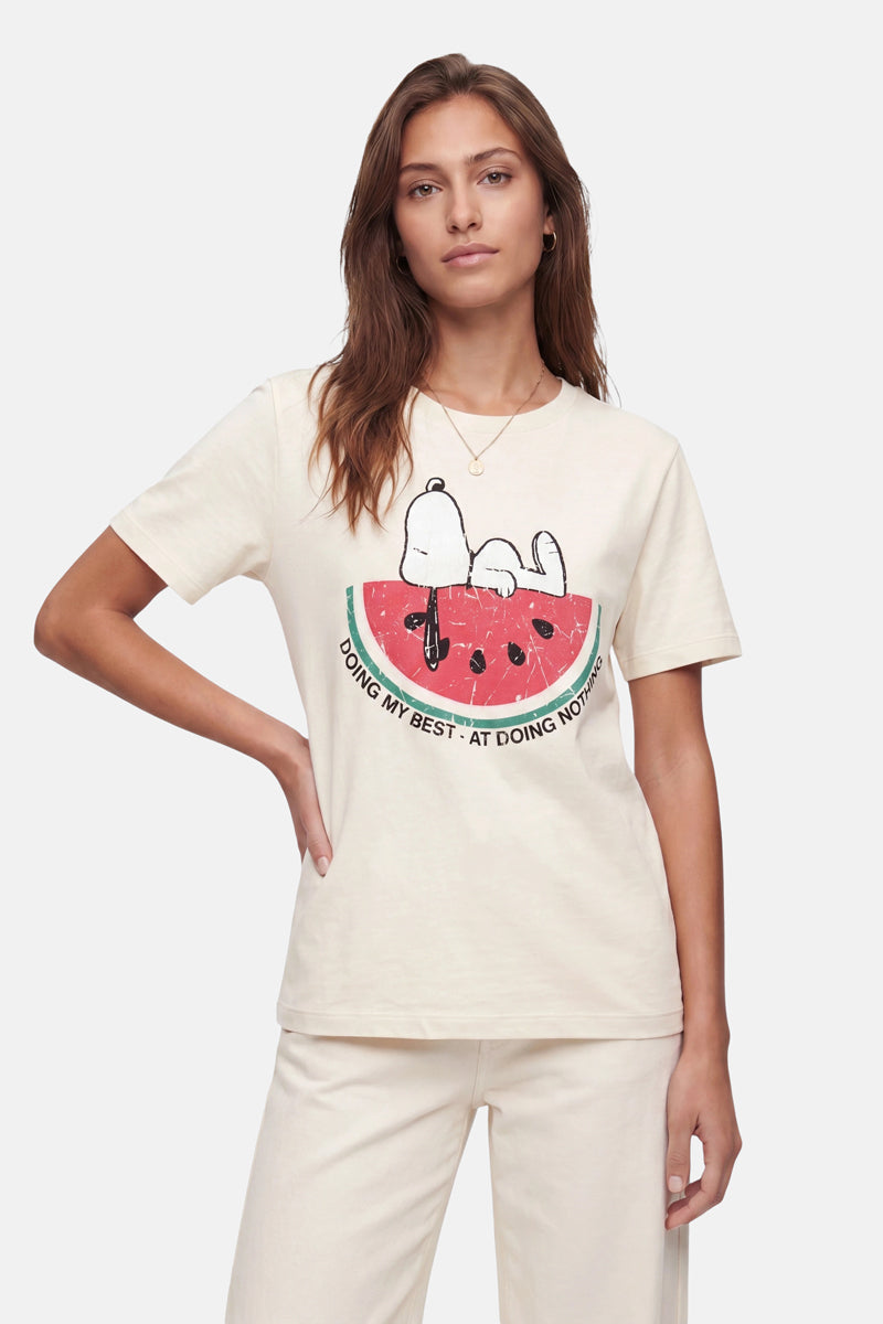 Eine Frau mit langen braunen Haaren trägt ein cremefarbenes Snoopy-T-Shirt der „Frogbox by Princess Limited Edition“ aus weicher Baumwolle, auf dem Snoopy auf einer Wassermelonenscheibe liegend zu sehen ist, zusammen mit dem Schriftzug „DOING MY BEST – AT DOING NOTHING“, dazu eine passende Hose.
