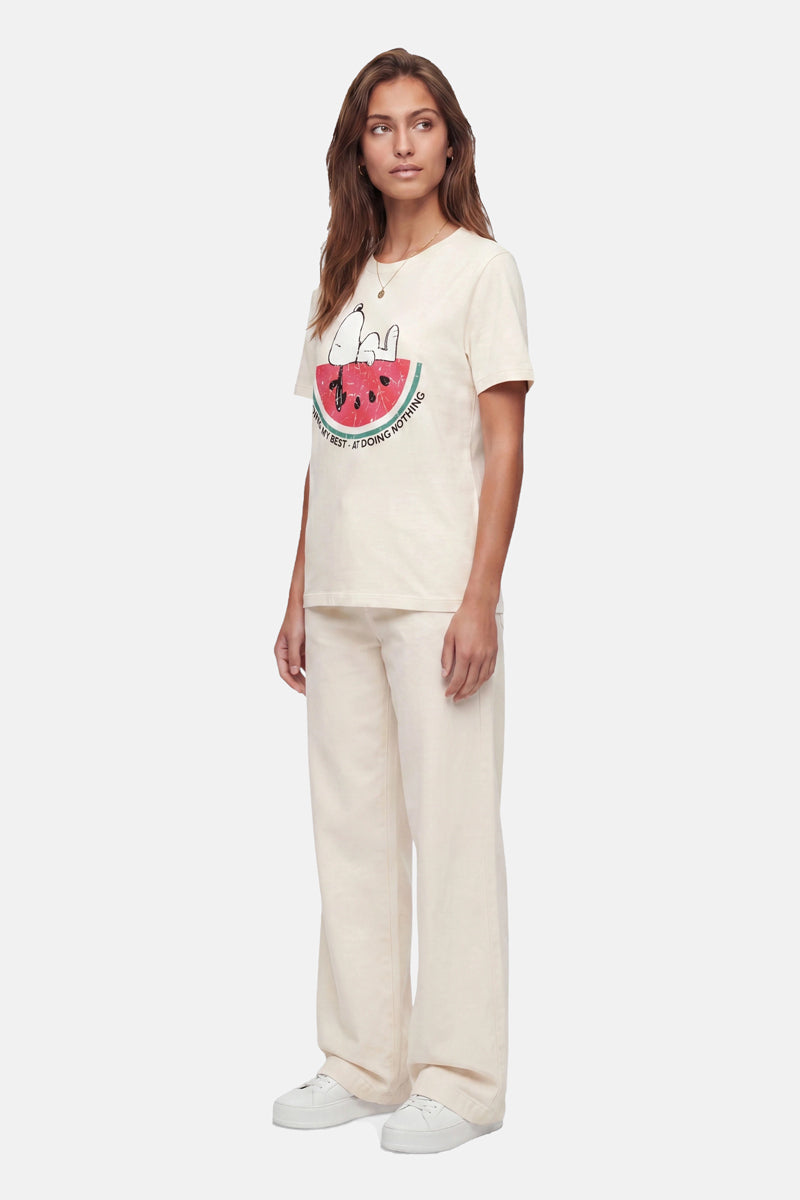 Vor einem hellgrauen Hintergrund steht eine Frau mit langen braunen Haaren und ausdruckslosem Gesicht. Sie trägt ein „Frogbox by Princess Limited Edition Snoopy“-T-Shirt aus Baumwolle, eine lockere, cremefarbene Hose und weiße Sneakers.
