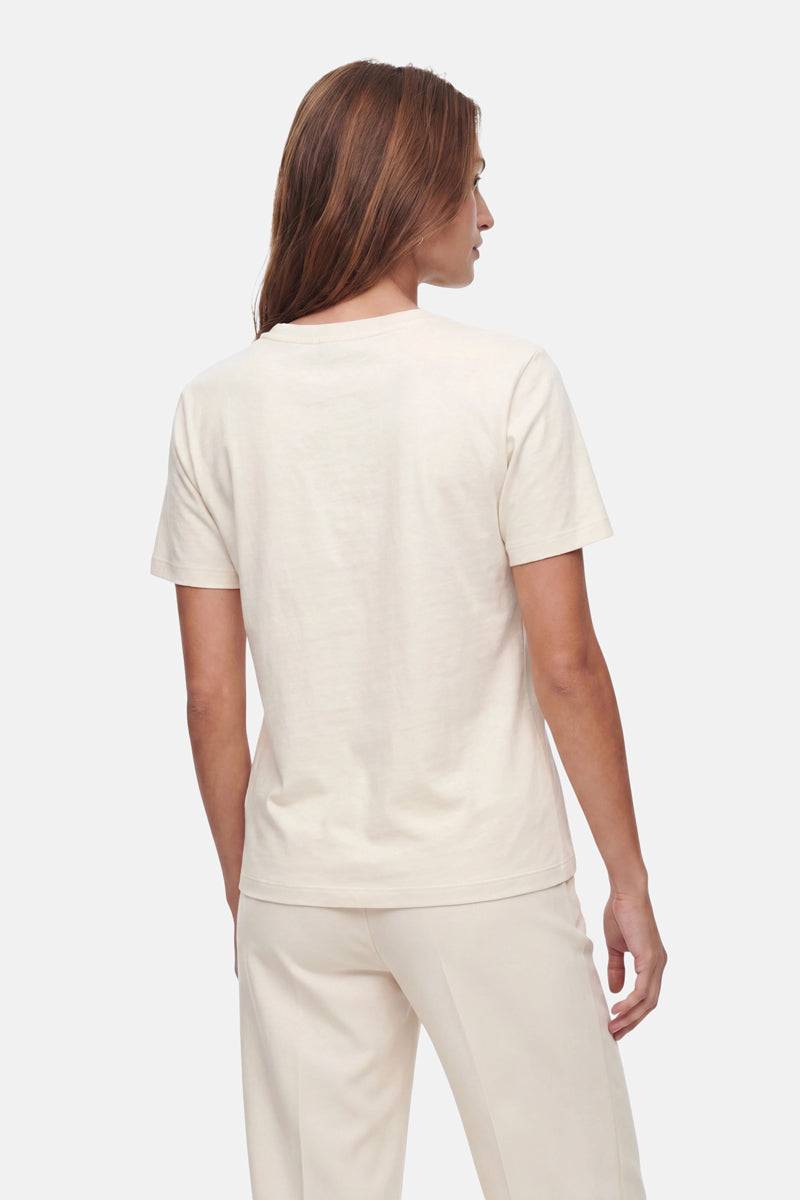 Eine Frau mit langen braunen Haaren steht mit dem Rücken zur Kamera und trägt ein cremefarbenes Snoopy-T-Shirt in limitierter Auflage sowie eine dazu passende Hose von „Frogbox by Princess“ vor einem schlichten hellgrauen Hintergrund.