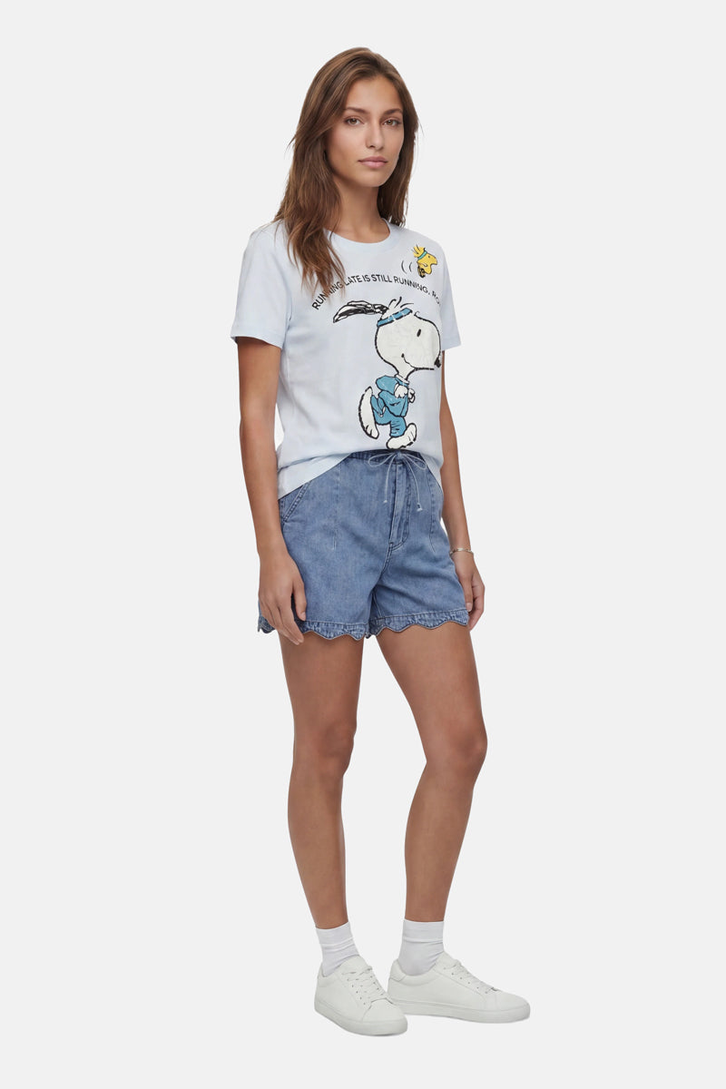 Eine Frau posiert lässig vor weißem Hintergrund in einem „Frogbox by Princess Limited Edition Snoopy“-T-Shirt aus Baumwolle, dazu trägt sie hellblaue Jeansshorts mit Wellenrand, weiße Söckchen und Sneakers; ihr Haar fällt ihr locker über die Schultern.