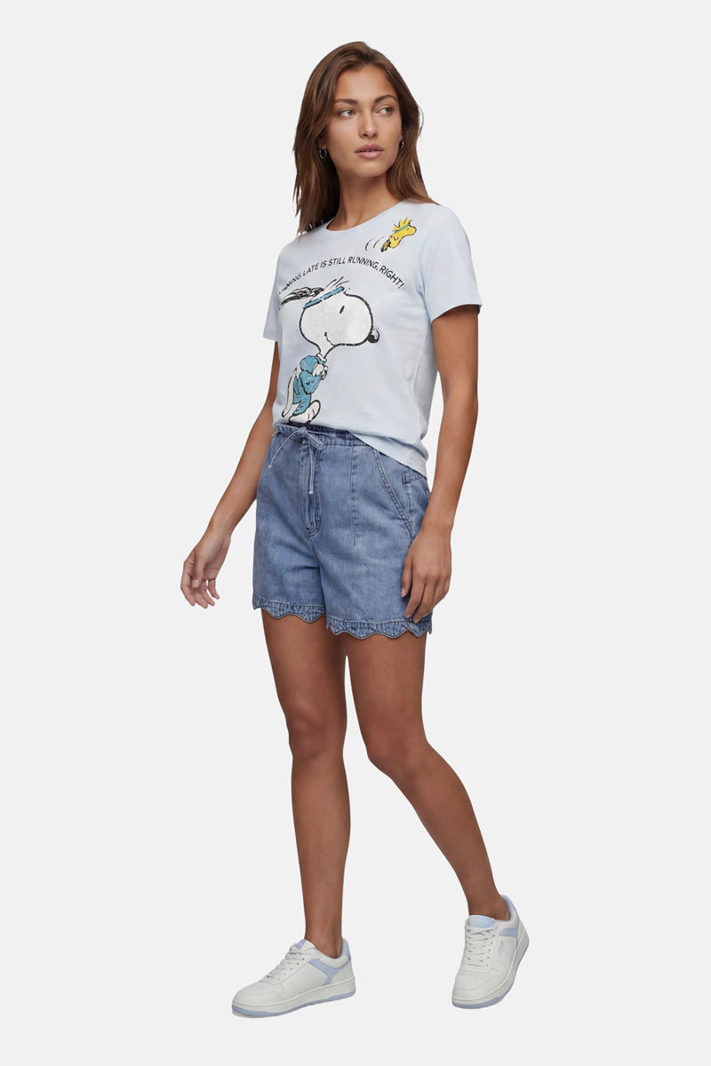 Vor einem schlichten hellen Hintergrund steht eine Frau mit langen braunen Haaren und einem entspannten Gesichtsausdruck. Sie trägt ein „Frogbox by Princess Limited Edition Snoopy“-T-Shirt aus Baumwolle, hellblaue Jeansshorts mit Wellenrand und weiße Sneakers.