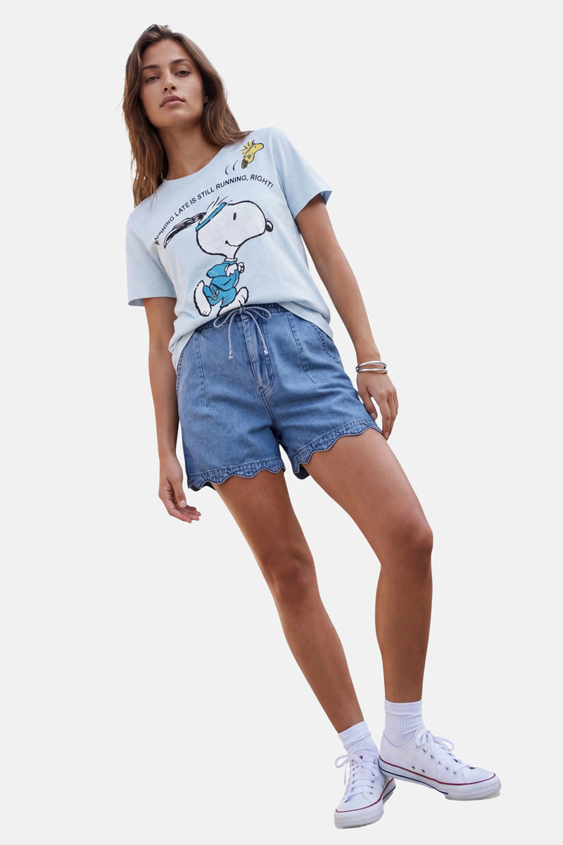 Eine junge Frau posiert vor einem weißen Hintergrund und trägt ein hellblaues Snoopy-T-Shirt aus Baumwolle der „Frogbox by Princess Limited Edition“, hoch taillierte Jeansshorts mit Wellenrand, weiße Söckchen und weiße High-Top-Sneaker; sie steht selbstbewusst da, ein Bein angewinkelt.