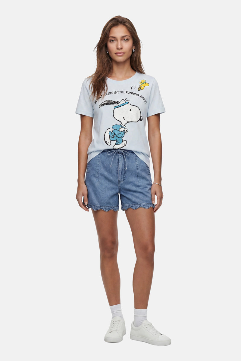 Eine Frau mit langen braunen Haaren blickt vor einem weißen Hintergrund nach vorne. Sie trägt ein hellblaues „Frogbox by Princess Limited Edition Snoopy“-T-Shirt aus Baumwolle, Jeans-Shorts mit Kordelzug und gewellten Säumen, weiße Söckchen und weiße Sneaker.