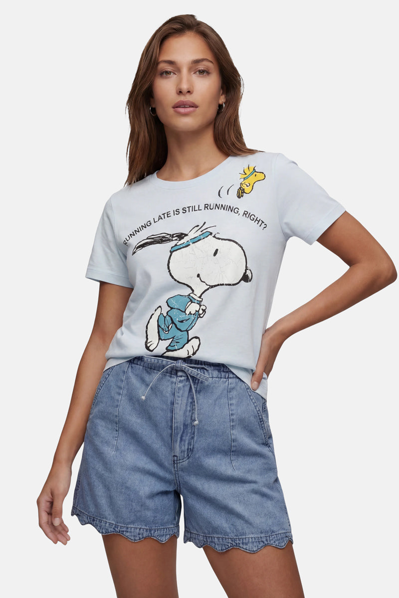 Eine Frau mit langen braunen Haaren trägt ein „Frogbox by Princess Limited Edition Snoopy“-T-Shirt aus weicher Baumwolle, auf dem Snoopy zusammen mit Woodstock zu sehen ist, sowie den Schriftzug „RUNNING LATE IS STILL RUNNING, RIGHT?“, dazu blaue Shorts mit Wellenrand.