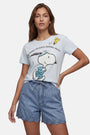 Eine Frau mit langen braunen Haaren trägt ein „Frogbox by Princess Limited Edition Snoopy“-T-Shirt aus weicher Baumwolle, auf dem Snoopy zusammen mit Woodstock zu sehen ist, sowie den Schriftzug „RUNNING LATE IS STILL RUNNING, RIGHT?“, dazu blaue Shorts mit Wellenrand.