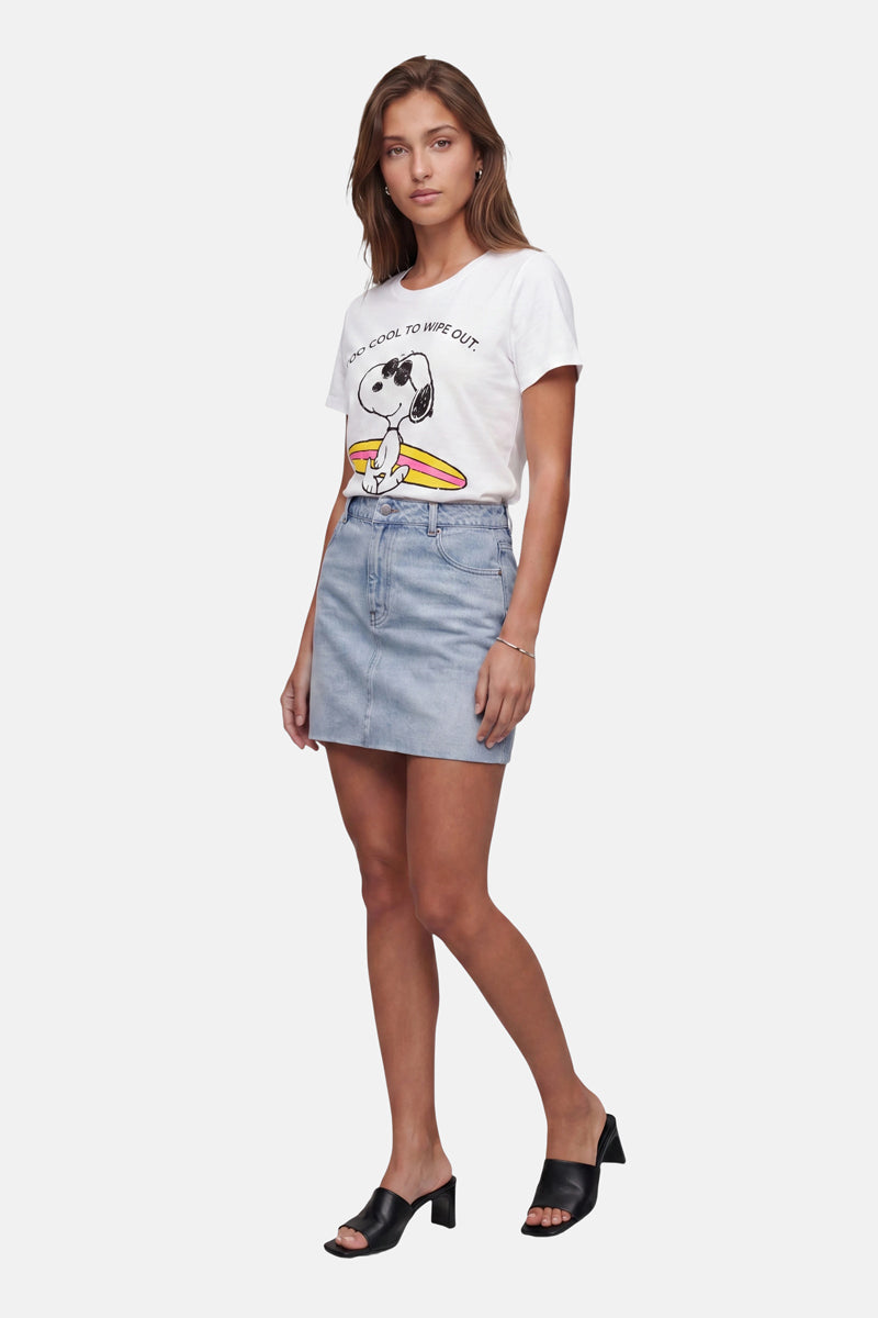 Eine Frau blickt nach vorne und trägt ein weißes Baumwoll-T-Shirt mit Snoopy-Aufdruck aus der limitierten „Frogbox by Princess“-Kollektion, einen hellblauen Jeansrock und schwarze Sandalen mit Absatz. Sie hat hellbraunes Haar, einen neutralen Gesichtsausdruck und hält ihre linke Hand an der Seite, vor einem schlichten weißen Hintergrund.