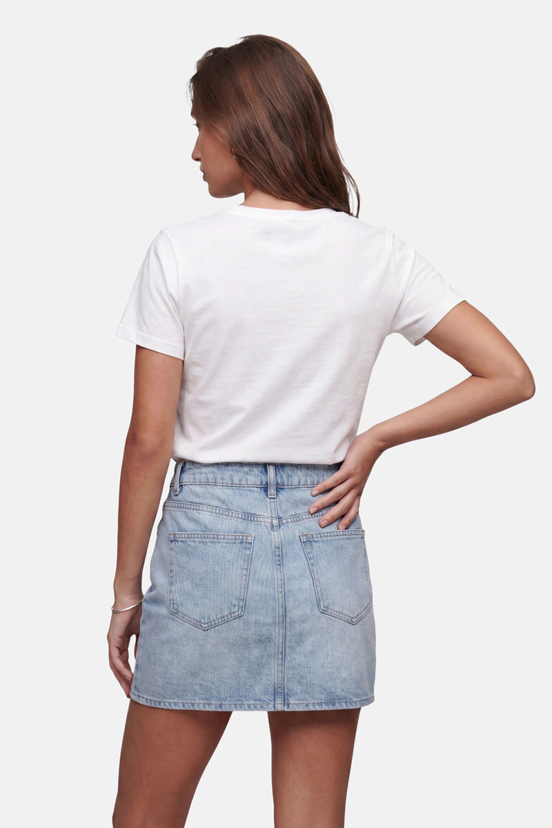 Eine Frau mit hellbraunen Haaren steht mit dem Rücken zur Kamera und trägt ein schlichtes weißes Baumwoll-T-Shirt der „Frogbox by Princess Limited Edition“, das in einen hellblauen Jeans-Minirock mit Gesäßtaschen gesteckt ist. Ihre rechte Hand ruht auf ihrer Hüfte vor einem schlichten weißen Hintergrund.