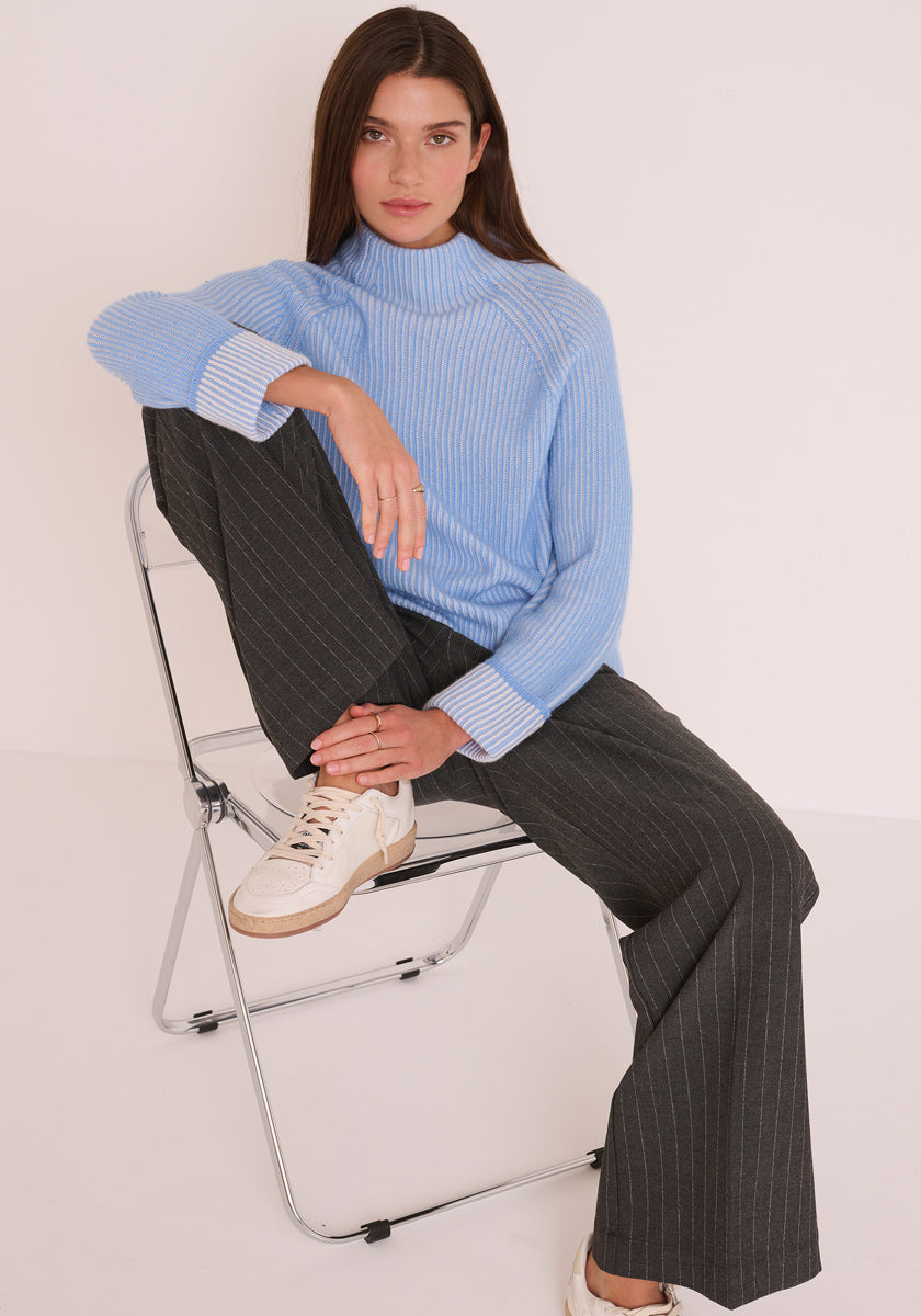 Eine Frau mit glattem braunem Haar sitzt auf einem Klappstuhl und trägt einen hellblauen Princess Goes Hollywood Rippstrickpullover aus Wolle-Kaschmir-Mix mit Stehkragen, eine dunkelgraue Nadelstreifenhose mit weitem Bein und weiße Turnschuhe. Ihr linker Arm ruht auf ihrem angehobenen Knie, während sie in die Kamera schaut.