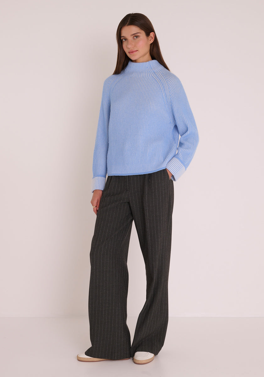 Eine Frau mit glattem braunem Haar posiert selbstbewusst vor einem schlichten Hintergrund. Sie trägt einen hellblauen Princess Goes Hollywood-Pullover aus einer Woll-Kaschmirmischung mit gerippten Bündchen und weißen Manschetten, eine dunkelgraue Nadelstreifenhose mit weitem Bein und weiße Turnschuhe.