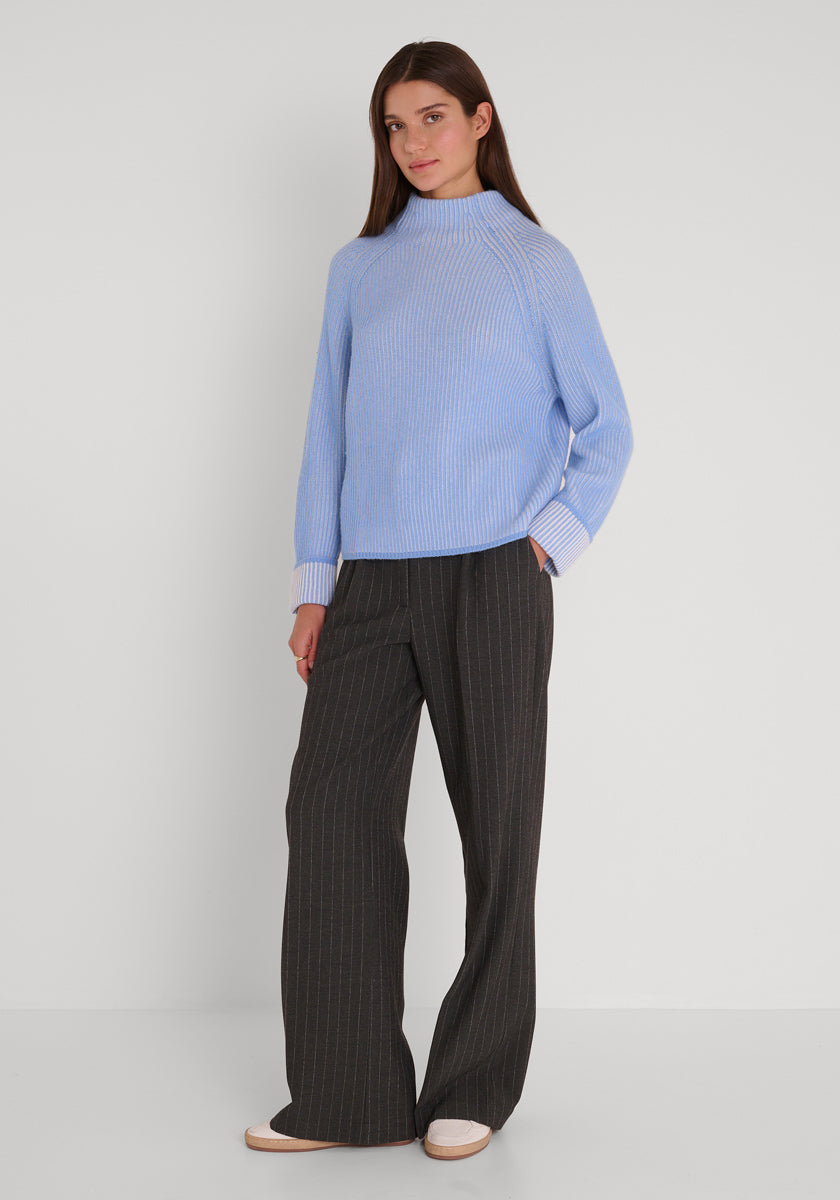 Eine Frau mit glattem braunem Haar trägt einen lockeren hellblauen Princess Goes Hollywood Rippstrickpullover aus Wolle-Kaschmir-Mix mit weißen Bündchen und eine dunkelbraune Nadelstreifenhose mit weitem Bein. Sie steht vor einem schlichten weißen Hintergrund und schaut sanft in die Kamera.