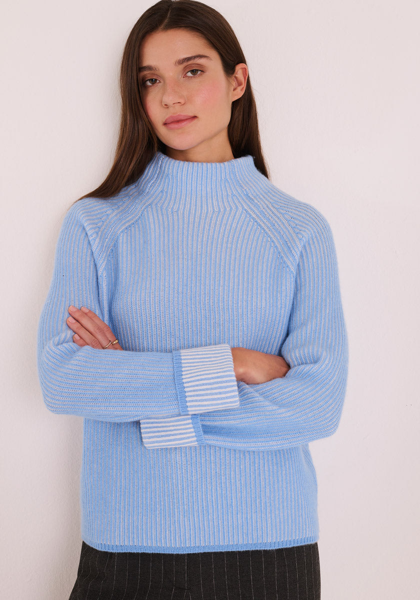 Eine Frau mit langen braunen Haaren trägt einen hellblauen Princess Goes Hollywood Pullover aus Woll-Kaschmir-Rippstrick mit umgeschlagenen Manschetten, die Streifen zeigen. Sie steht mit verschränkten Armen vor einer weißen Wand und kombiniert ihn mit einer dunklen Hose oder einem Rock.