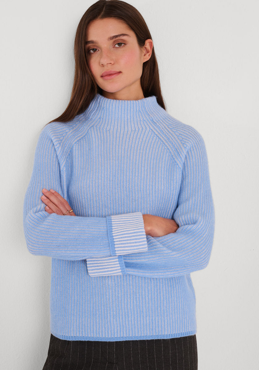 Eine Frau mit langen braunen Haaren steht mit verschränkten Armen vor einer weißen Wand. Sie trägt einen hellblauen Rippstrickpullover mit Stehkragen aus Wolle-Kaschmir-Mix und dunkle Nadelstreifenhosen von Princess Goes Hollywood. Ihr Blick ist neutral.