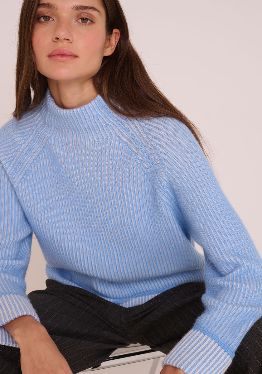 Eine Frau mit langen braunen Haaren trägt einen hellblauen Princess Goes Hollywood Woll-Kaschmir-Rippenpullover mit Stehkragen und eine dunkle Nadelstreifenhose. Sie sitzt auf einem Hocker, blickt nach vorne und hat einen neutralen Gesichtsausdruck vor einem schlichten hellen Hintergrund.