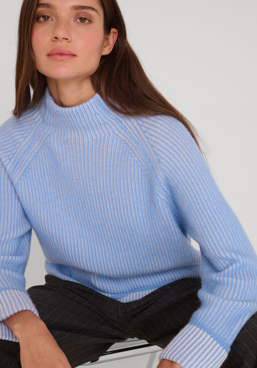 Eine Frau mit langen braunen Haaren sitzt auf einem Hocker vor weißem Hintergrund und trägt einen hellblauen Princess Goes Hollywood Rippstrickpullover aus Wolle-Kaschmir-Mix mit Stehkragen und weiten Ärmeln, gepaart mit einer dunklen Nadelstreifhose. Sie blickt ruhig in die Kamera.