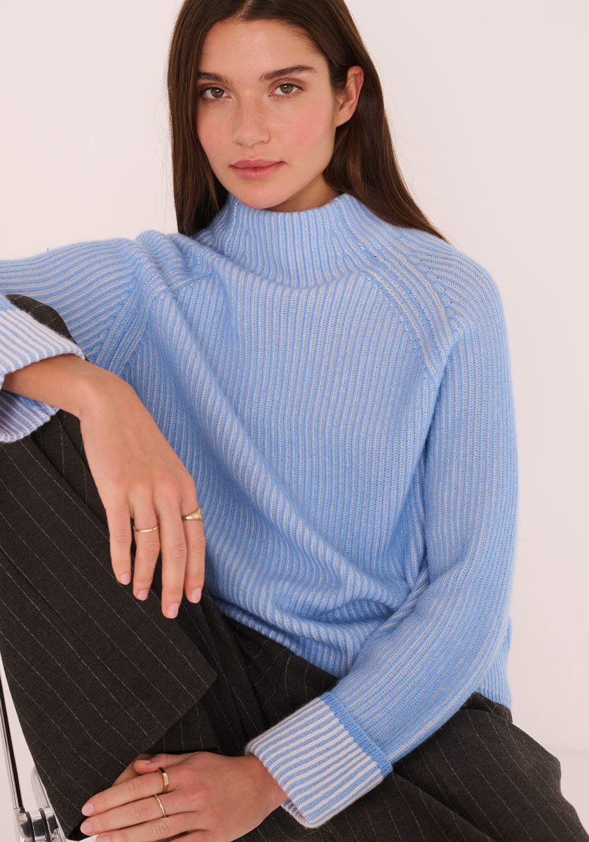 Eine Frau mit langen braunen Haaren und minimalem Schmuck sitzt mit einem Knie hoch und trägt einen hellblauen Princess Goes Hollywood Rollkragenpullover aus Wolle und Kaschmir und eine graue Nadelstreifenhose vor einem schlichten hellen Hintergrund.