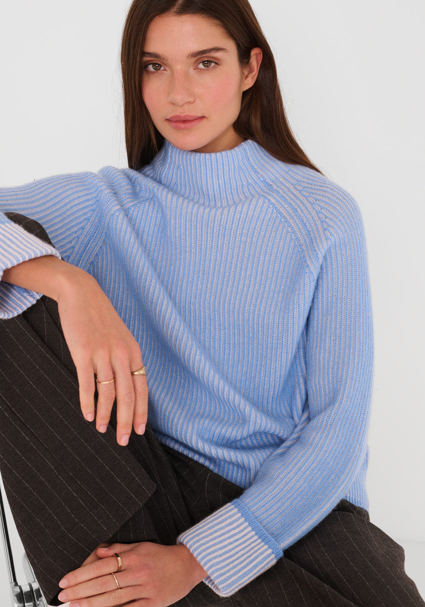 Eine Frau mit langen braunen Haaren trägt einen himmelblauen und weißen Princess Goes Hollywood Woll-Kaschmir-Rippenpullover mit Stehkragen, dazu eine braune Nadelstreifenhose. Sie sitzt ruhig da, ein Knie hoch, Goldringe an den Fingern.