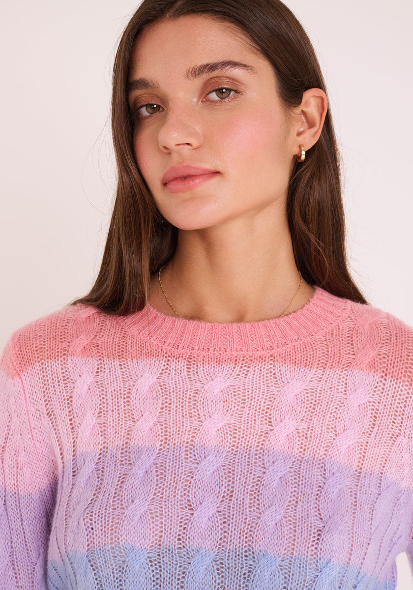 Eine junge Frau mit langen braunen Haaren trägt einen pastellrosa und lilafarbenen Princess Goes Hollywood Zopfstrickpullover aus Merino-Alpaka-Mix mit Rundhalsausschnitt und kleinen goldenen Reifohrringen. Sie blickt leicht zur Seite, neutraler Ausdruck, vor einem schlichten hellen Hintergrund.