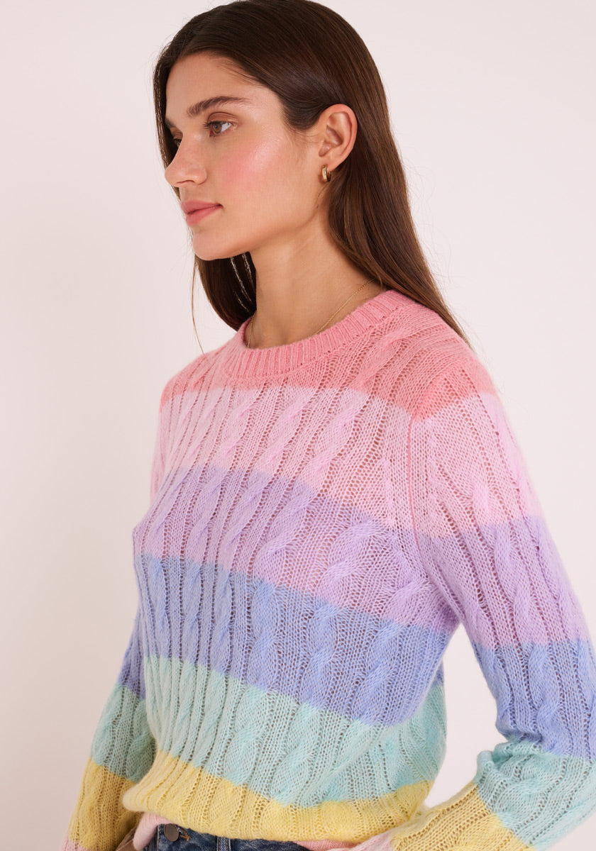 Eine Frau mit langen braunen Haaren trägt einen pastellfarbenen regenbogengestreiften Zopfstrickpullover aus Merino-Alpaka-Mix mit Rundhalsausschnitt von Princess Goes Hollywood. Sie steht links im Seitenprofil vor einem hellen Hintergrund, ihr Gesichtsausdruck ist neutral.