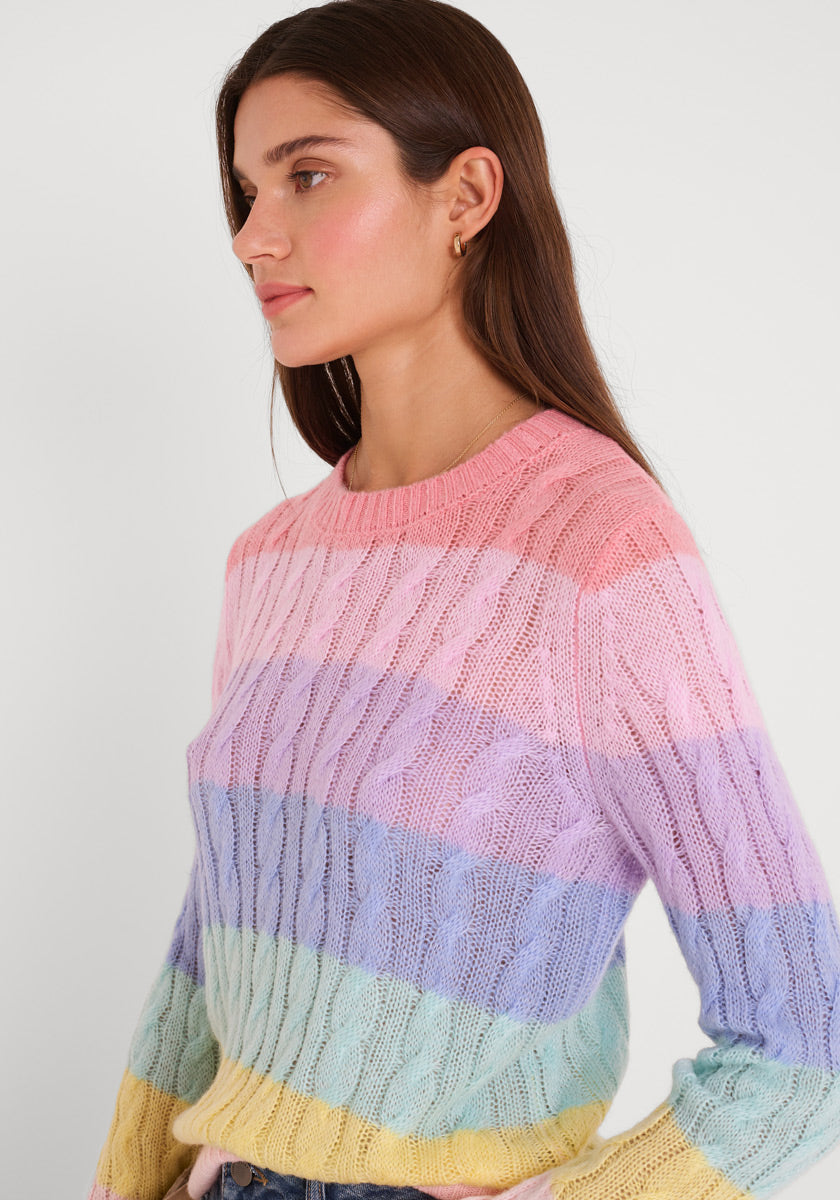 Eine Frau mit langen braunen Haaren und kleinen Ohrringen trägt einen Princess Goes Hollywood gestreifter Zopfstrickpullover in Pastellrosa, Lila, Blau und Gelb. Sie blickt nach links vor einem schlichten weißen Hintergrund und hat einen ruhigen Gesichtsausdruck.