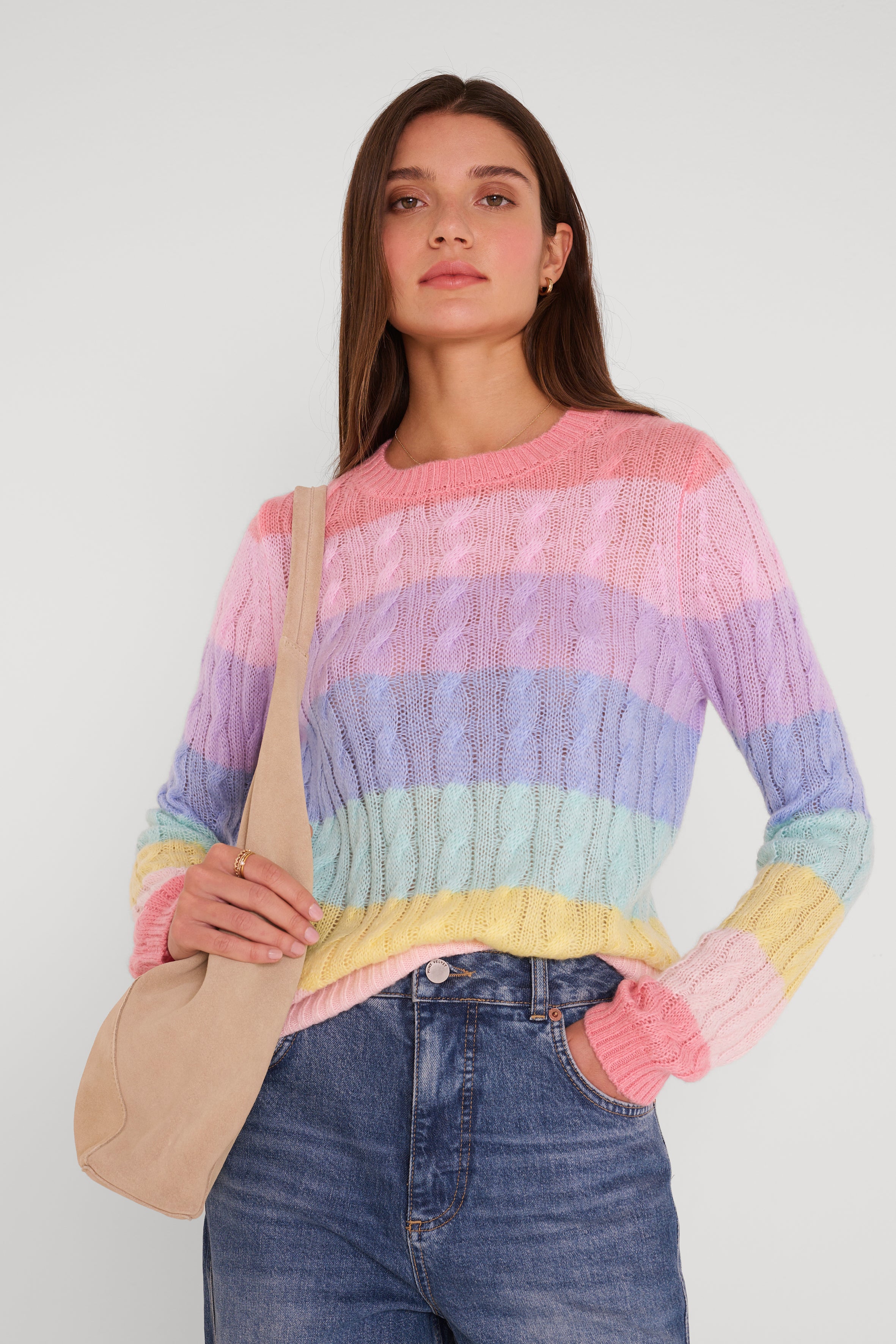 Eine Frau mit glattem braunem Haar und neutralem Gesichtsausdruck steht vor einem schlichten Hintergrund. Sie trägt eine blaue Jeans und einen pastellfarben gestreiften Pullover aus einem Merino-Alpaka-Mix von Princess Goes Hollywood. Sie trägt eine beigefarbene Umhängetasche, die Hand ruht auf dem Trageriemen.