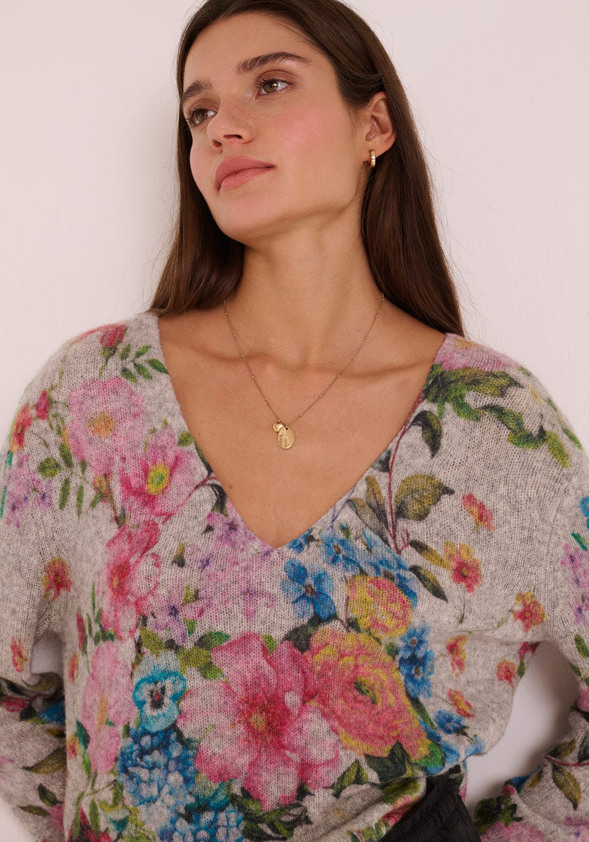 Eine Frau mit langen braunen Haaren trägt einen grauen Princess Goes Hollywood Strickpullover aus Wollmix mit buntem Blumen-Print in Pink, Blau und Gelb. Sie trägt goldene Accessoires und blickt vor weißem Hintergrund leicht nach oben.