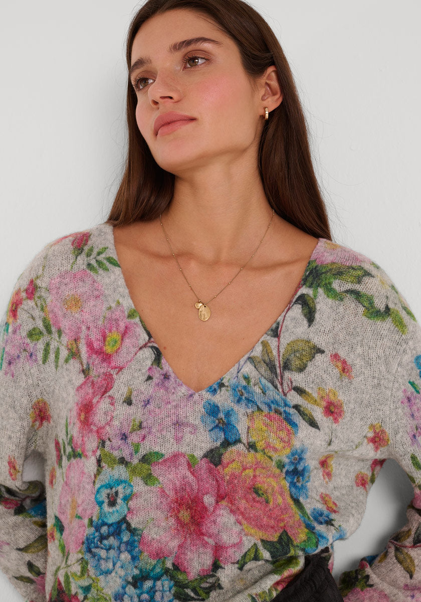 Eine Frau mit langen braunen Haaren trägt einen hellgrauen Princess Goes Hollywood Strickpullover aus Wollmix mit leuchtendem rosa, blauen und grünen Blumendruck. Dazu trägt sie eine goldene Halskette und kleine goldene Reifen vor einem schlichten weißen Hintergrund.