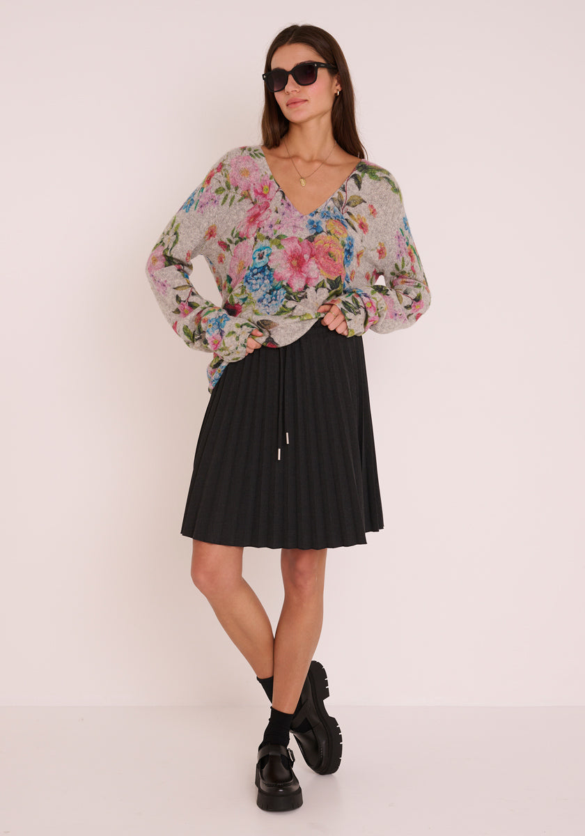 Eine Frau steht vor einem schlichten Hintergrund und trägt eine dunkle Sonnenbrille, einen bunten, geblümten Wollmix Strickpullover von Princess Goes Hollywood, einen schwarzen Faltenrock, schwarze Socken und klobige schwarze Schuhe. Ihre Hände stecken in den Taschen ihres Pullovers und sie sieht entspannt aus.