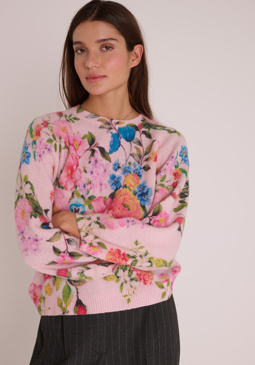 Eine Frau mit langen dunklen Haaren trägt einen hellrosa Princess Goes Hollywood Strickpullover aus Wollmix mit einem bunten blauen, rosa und gelben Blumenprint. Sie steht vor einem schlichten hellen Hintergrund, die Arme verschränkt, in einer dunklen Hose.