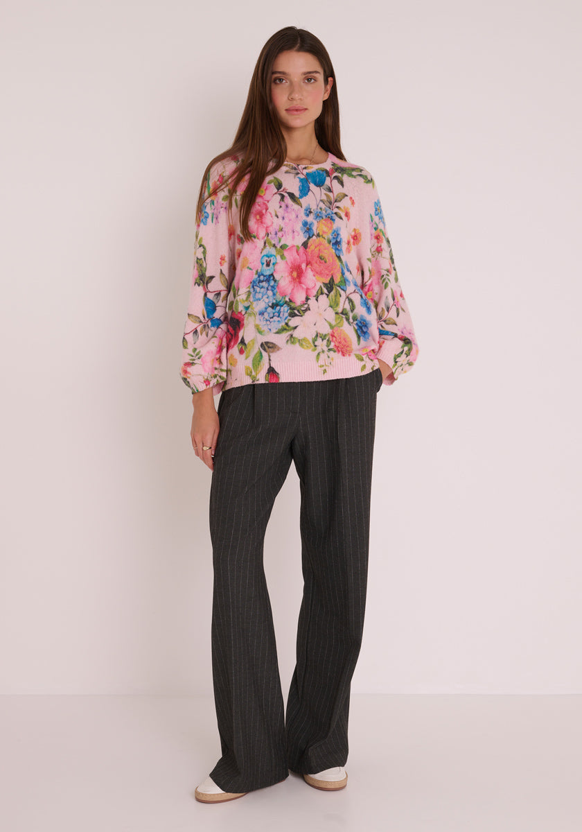 Eine Frau mit langen braunen Haaren steht vor einer weißen Wand. Sie trägt einen lockeren rosafarbenen Blumen-Print Wollmix Strickpullover und eine locker sitzende dunkelgraue Nadelstreifenhose von Princess Goes Hollywood. Sie hat einen neutralen Gesichtsausdruck und helle Schuhe.