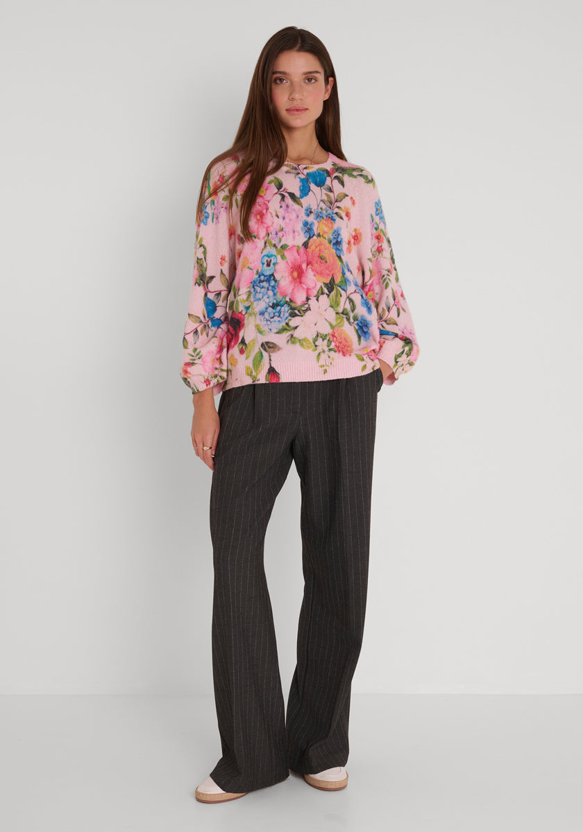 Eine Frau mit langen braunen Haaren steht vor einer weißen Wand. Sie trägt einen lockeren hellrosa Princess Goes Hollywood Strickpullover mit Blumenaufdruck, eine dunkle Hose mit weitem Bein in Nadelstreifen und dazu passende hellrosa Schuhe. Ihre Arme ruhen entspannt an ihren Seiten.