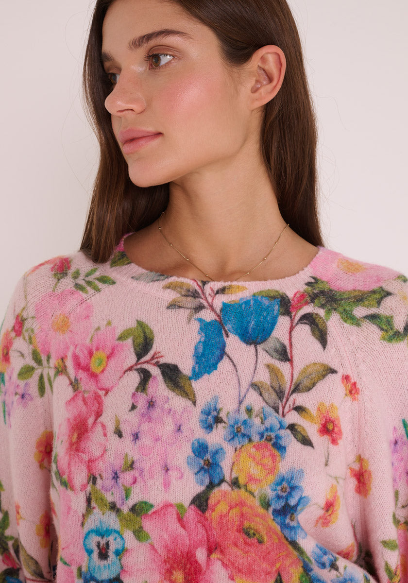 Eine Frau mit langen braunen Haaren blickt nach links und trägt einen hellrosa Princess Goes Hollywood Wollmix Strickpullover mit einem bunten Blumenmuster in Blau, Gelb und Rosa. Ihre Haut leuchtet natürlich und sie trägt eine zarte goldene Halskette.