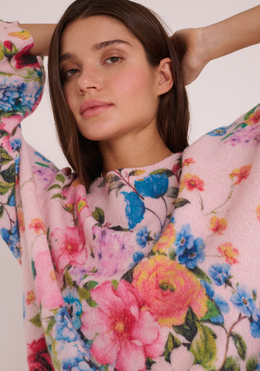 Eine junge Frau mit glattem braunem Haar posiert selbstbewusst, die Hände hinter dem Kopf, in einem zartrosa Princess Goes Hollywood Wollmix Strickpullover, der mit einem leuchtend blauen, rosa, gelben und grünen Blumen-Print verziert ist.