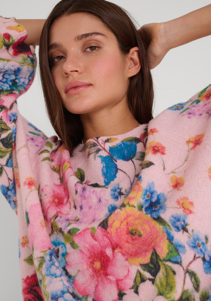 Eine Frau mit langen braunen Haaren blickt nach vorne, die Arme hinter dem Kopf verschränkt, und trägt einen zartrosa Princess Goes Hollywood Strickpullover mit leuchtend blauem, gelbem, rosa und grünem Blumendruck auf einfarbigem Hintergrund.