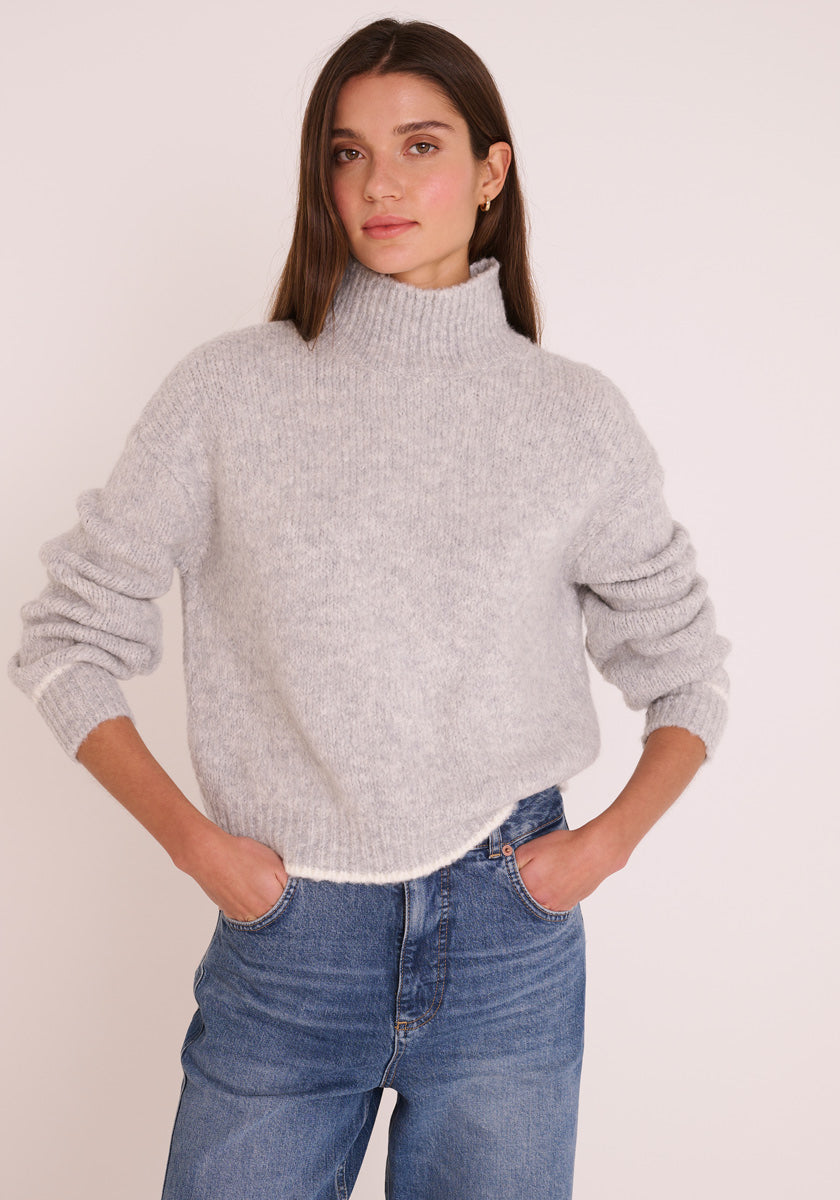 Eine Frau mit glattem braunem Haar trägt einen hellgrauen Princess Goes Hollywood Grobstrickpullover mit Stehkragen und langen Ärmeln und blaue Jeans. Sie steht mit den Händen in den Taschen vor einem schlichten Hintergrund und schaut leicht zur Seite.