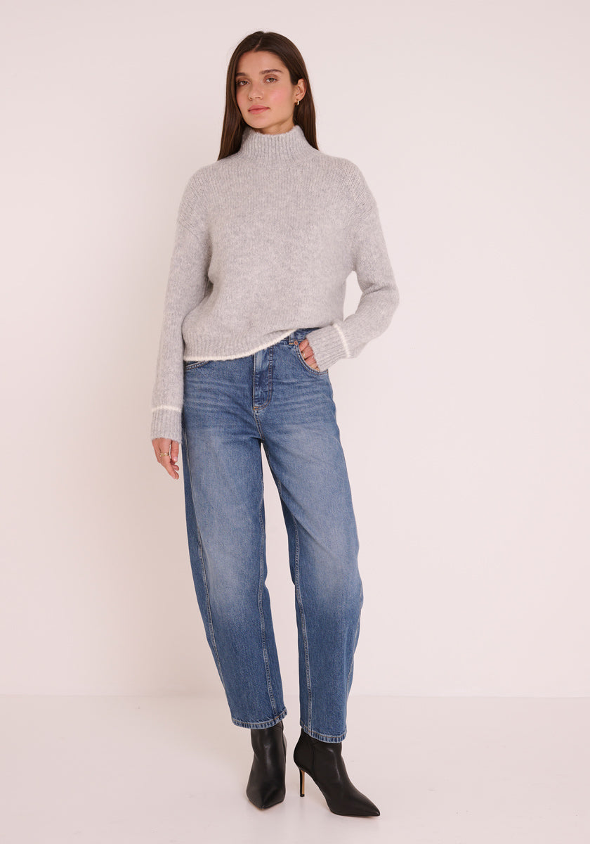 Eine Frau mit langen braunen Haaren trägt einen hellgrauen Princess Goes Hollywood Damen Pullover mit Stehkragen, lässige Bluejeans und schwarze spitze Ankle Boots. Sie steht vor einem weißen Hintergrund, hat eine Hand in der Tasche und einen neutralen Gesichtsausdruck.