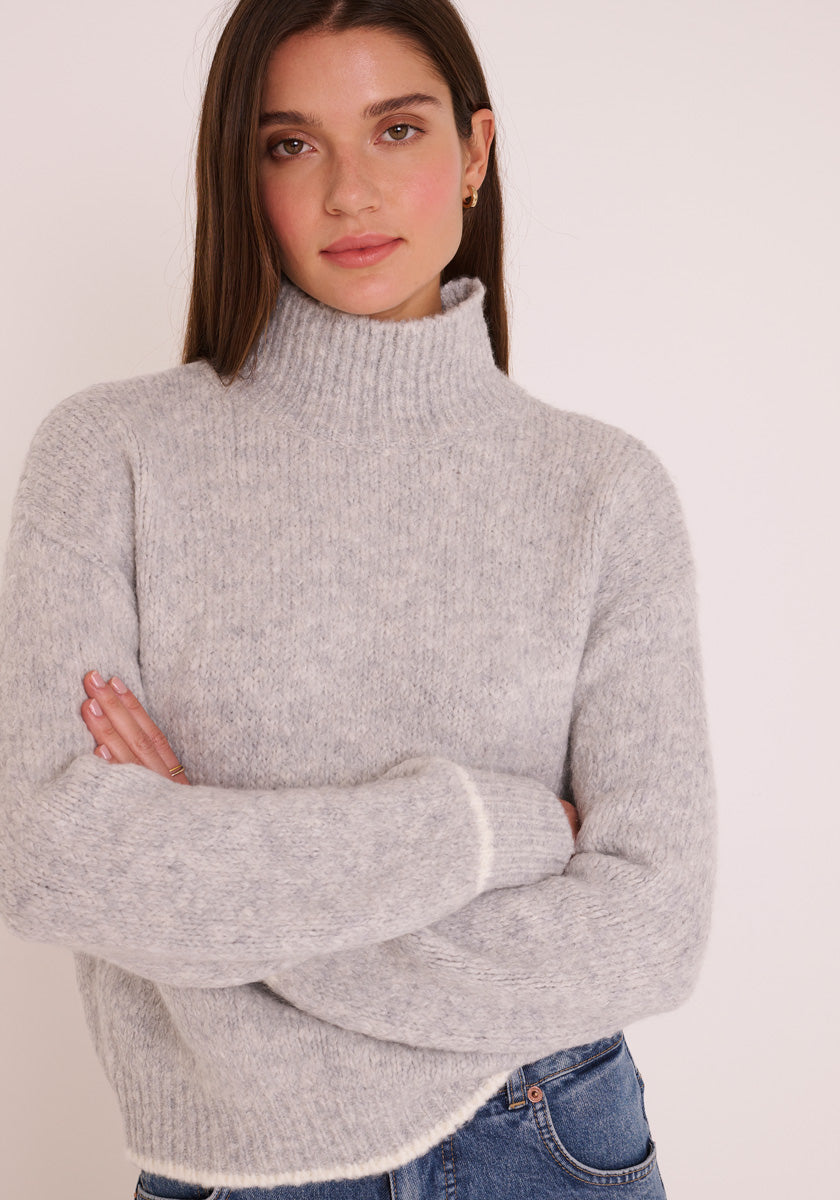 Eine Frau mit glattem braunem Haar trägt einen hellgrauen Princess Goes Hollywood Grobstrickpullover mit Stehkragen und langen Ärmeln, gepaart mit hochgekrempelten Blue Jeans. Sie steht mit verschränkten Armen vor einem schlichten hellen Hintergrund, ihr Gesichtsausdruck ist neutral.