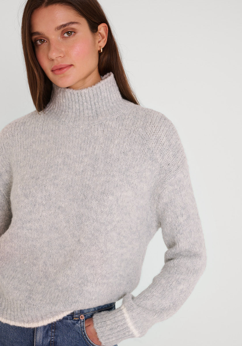 Eine Frau mit glattem braunem Haar trägt einen hellgrauen Damen Pullover mit Stehkragen von Princess Goes Hollywood und blaue Jeans. Sie steht vor einem weißen Hintergrund und blickt mit neutraler Miene zur Seite, die linke Hand in die Hüfte gestemmt.