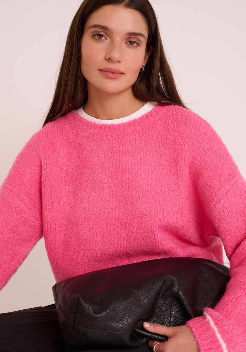 Eine Frau mit langen braunen Haaren trägt einen wuscheligen, hellrosa Princess Goes Hollywood Damen Pullover mit Rundhalsausschnitt über einem weißen Hemd, hält eine große schwarze Lederclutch im Schoß und sitzt vor einem hellen Hintergrund.