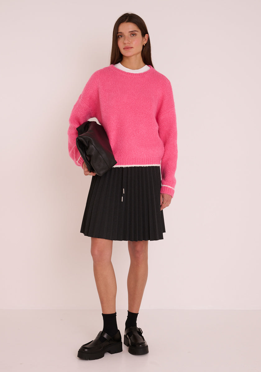 Eine Frau trägt einen leuchtend rosa Princess Goes Hollywood Grobstrickpullover mit langen Ärmeln und Rundhalsausschnitt über einem weißen Oberteil, einen schwarzen Faltenrock, Socken, klobige Schuhe und hält eine schwarze Clutch vor einem weißen Hintergrund.