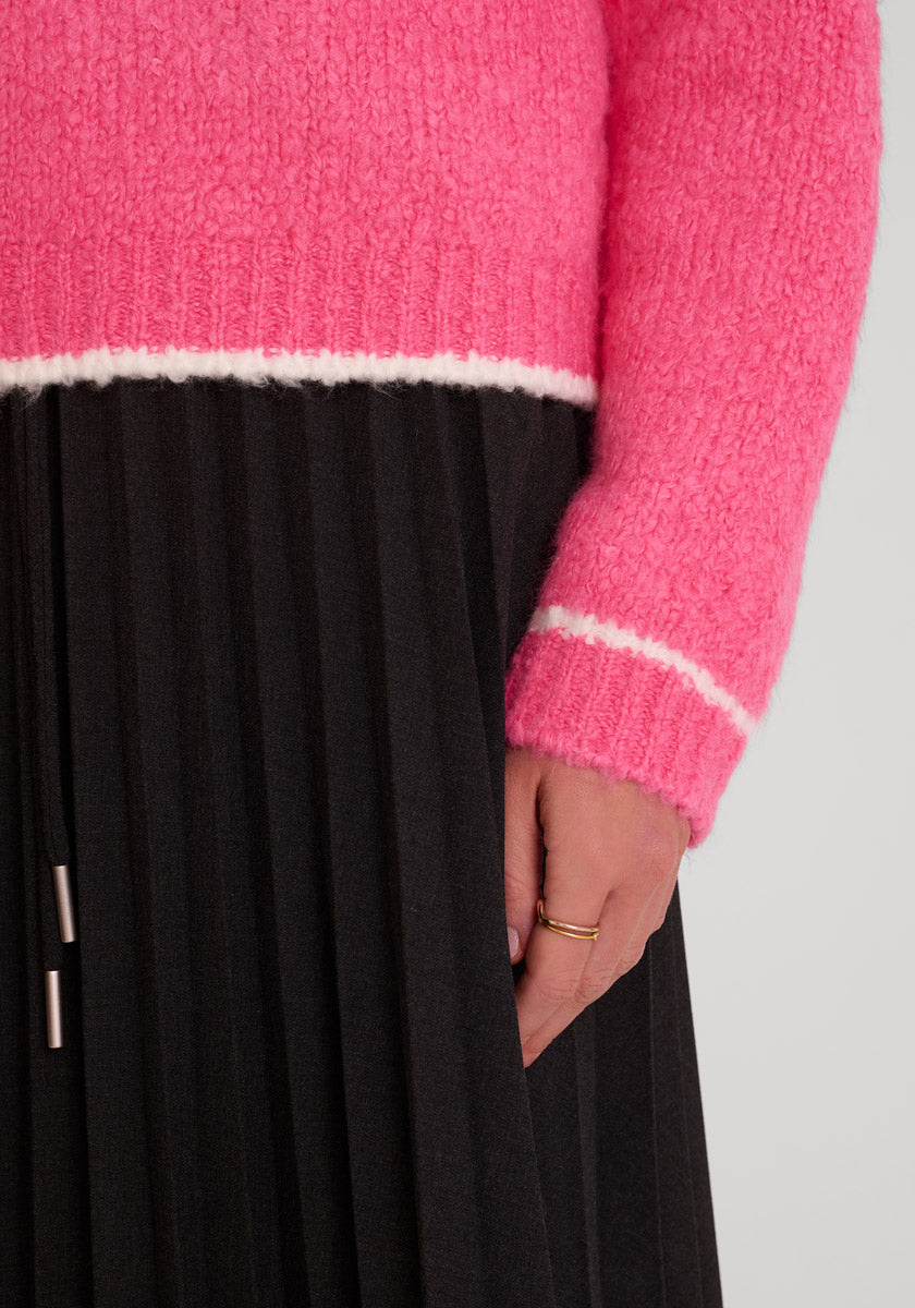 Eine Person trägt einen leuchtend rosafarbenen Princess Goes Hollywood Grobstrickpullover mit langen Ärmeln und weißen Streifen in der Nähe der Manschetten und des Saums, gepaart mit dünnen goldenen Ringen und einem schwarzen Faltenrock, vor einem schlichten hellgrauen Hintergrund.