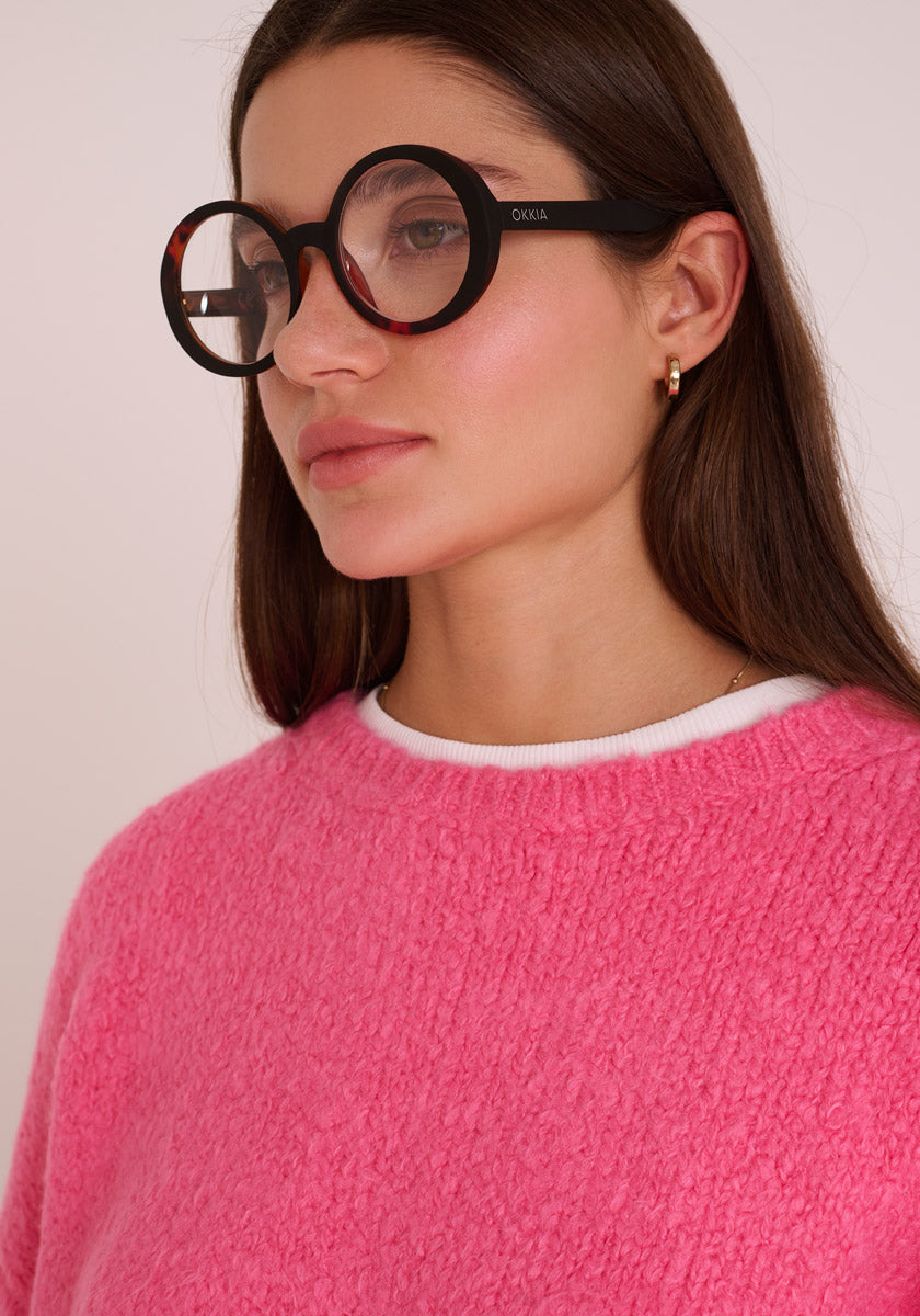 Eine Frau mit langen braunen Haaren und einer Schildpattbrille trägt kleine goldene Reifchen und einen leuchtend rosa Princess Goes Hollywood Grobstrickpullover mit Rundhalsausschnitt über einem weißen Oberteil vor einem hellen Hintergrund.