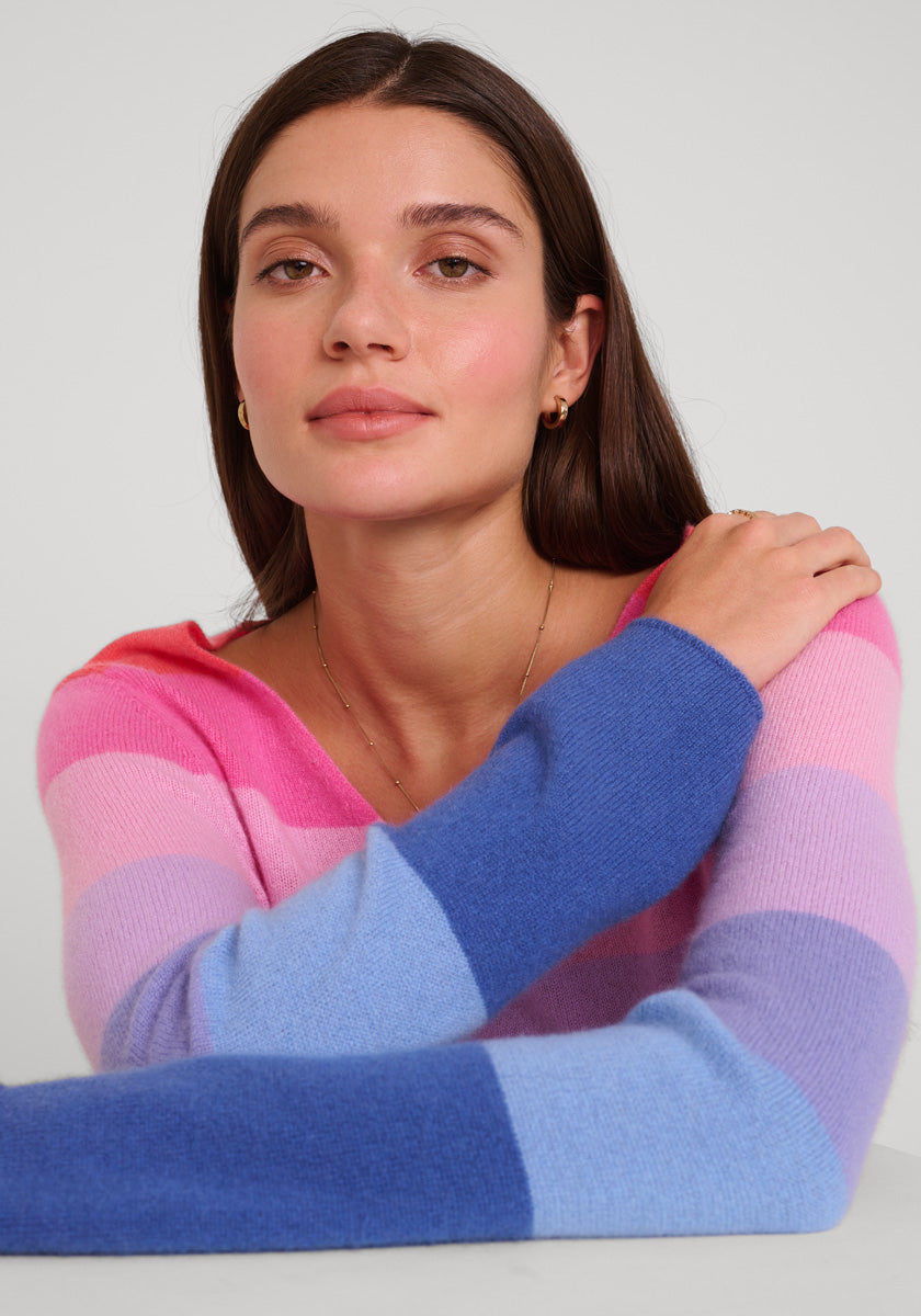 Eine Frau mit langen braunen Haaren und heller Haut sitzt an einem Tisch und schaut ruhig in die Kamera. Sie trägt einen gestreiften Princess Goes Hollywood Feinstrickpullover in Rosa, Lila und Blau vor einem schlichten Hintergrund.