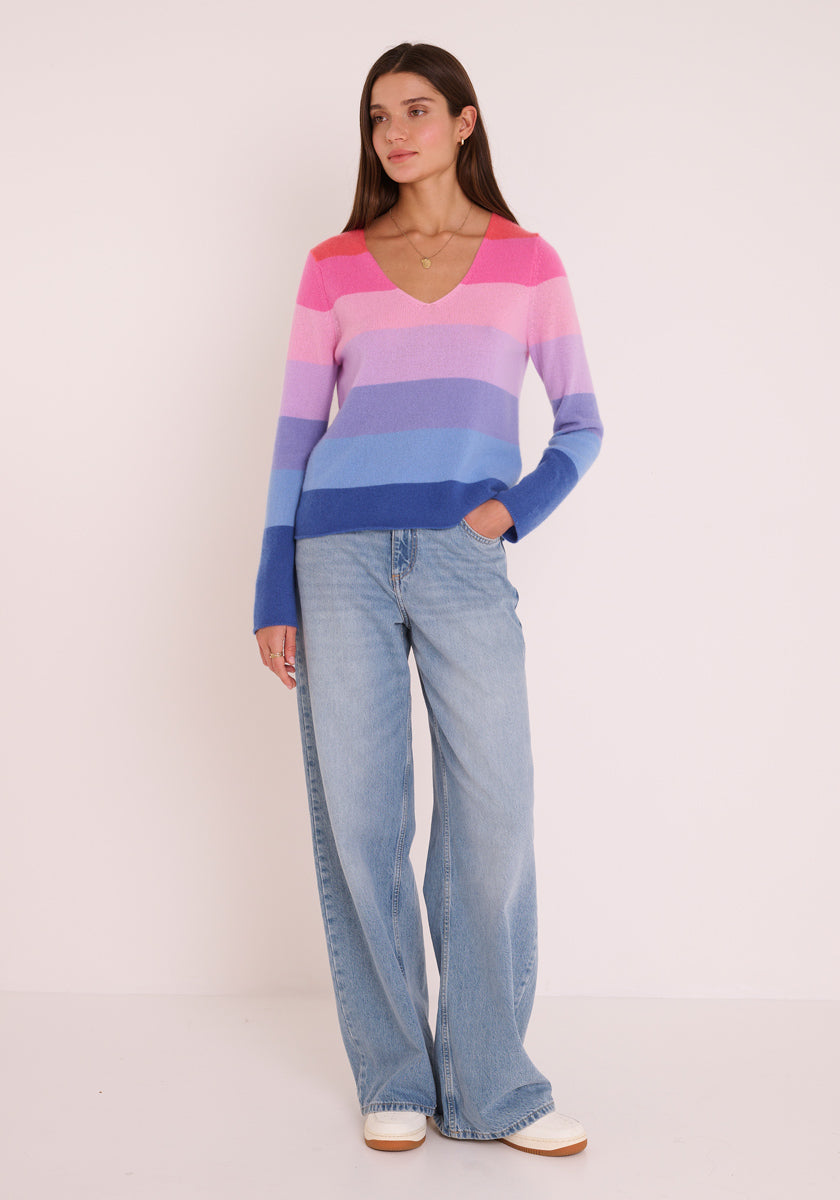 Eine Frau mit glattem braunem Haar und neutralem Gesichtsausdruck modelliert einen rosa, lila und blau gestreiften Feinstrickpullover mit V-Ausschnitt und langen Ärmeln, hellblaue Jeans mit weitem Bein und weiße Schuhe vor einem schlichten weißen Hintergrund.