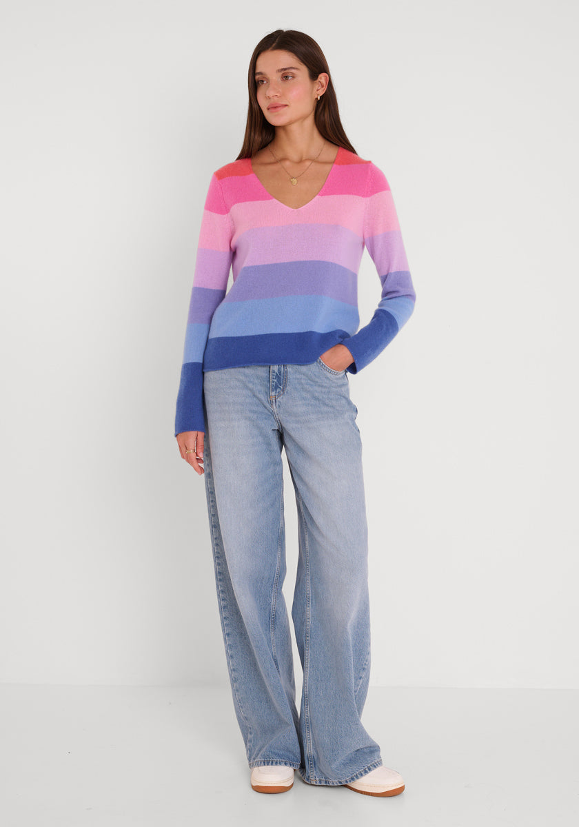 Eine Frau mit glattem braunem Haar trägt einen rosa, lila und blau gestreiften Feinstrickpullover mit V-Ausschnitt von Princess Goes Hollywood, hellblaue Jeans mit weitem Bein und weiße Schuhe. Sie posiert entspannt mit einer Hand in der Tasche und einer einfachen Halskette vor einem weißen Hintergrund.