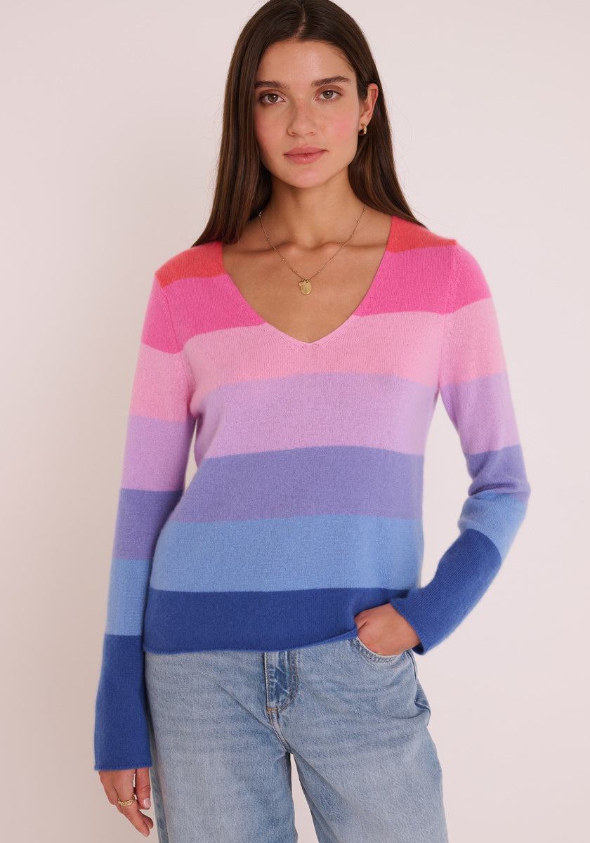 Eine Frau mit langen braunen Haaren trägt einen Feinstrickpullover mit V-Ausschnitt von Princess Goes Hollywood mit rosa, violetten und blauen Streifen, dazu eine hellgewaschene Jeans. Sie steht vor einem schlichten hellen Hintergrund, eine Hand in der Tasche.