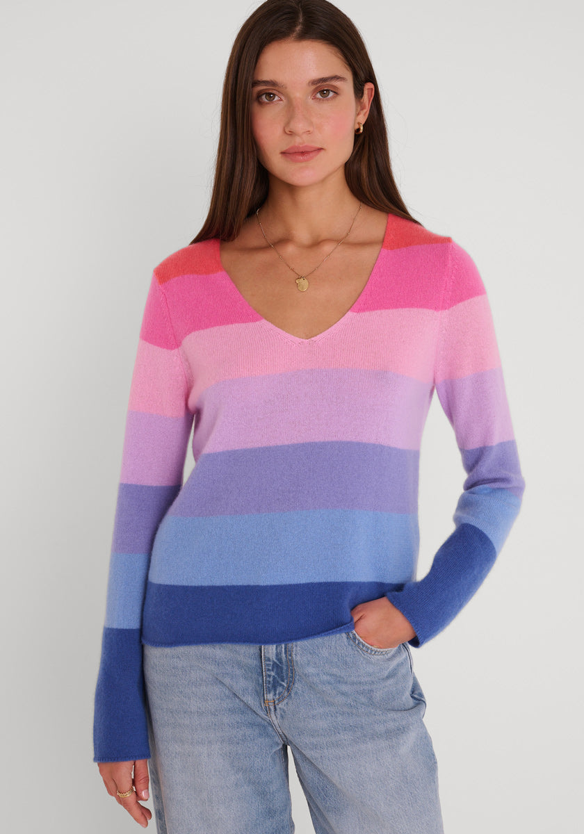Eine Frau mit langen braunen Haaren trägt einen Princess Goes Hollywood-Pullover mit V-Ausschnitt und breiten Querstreifen in Rosa, Lila und Blau. Sie steht vor einem schlichten hellen Hintergrund, eine Hand in der Jeanstasche, und schaut in die Kamera.